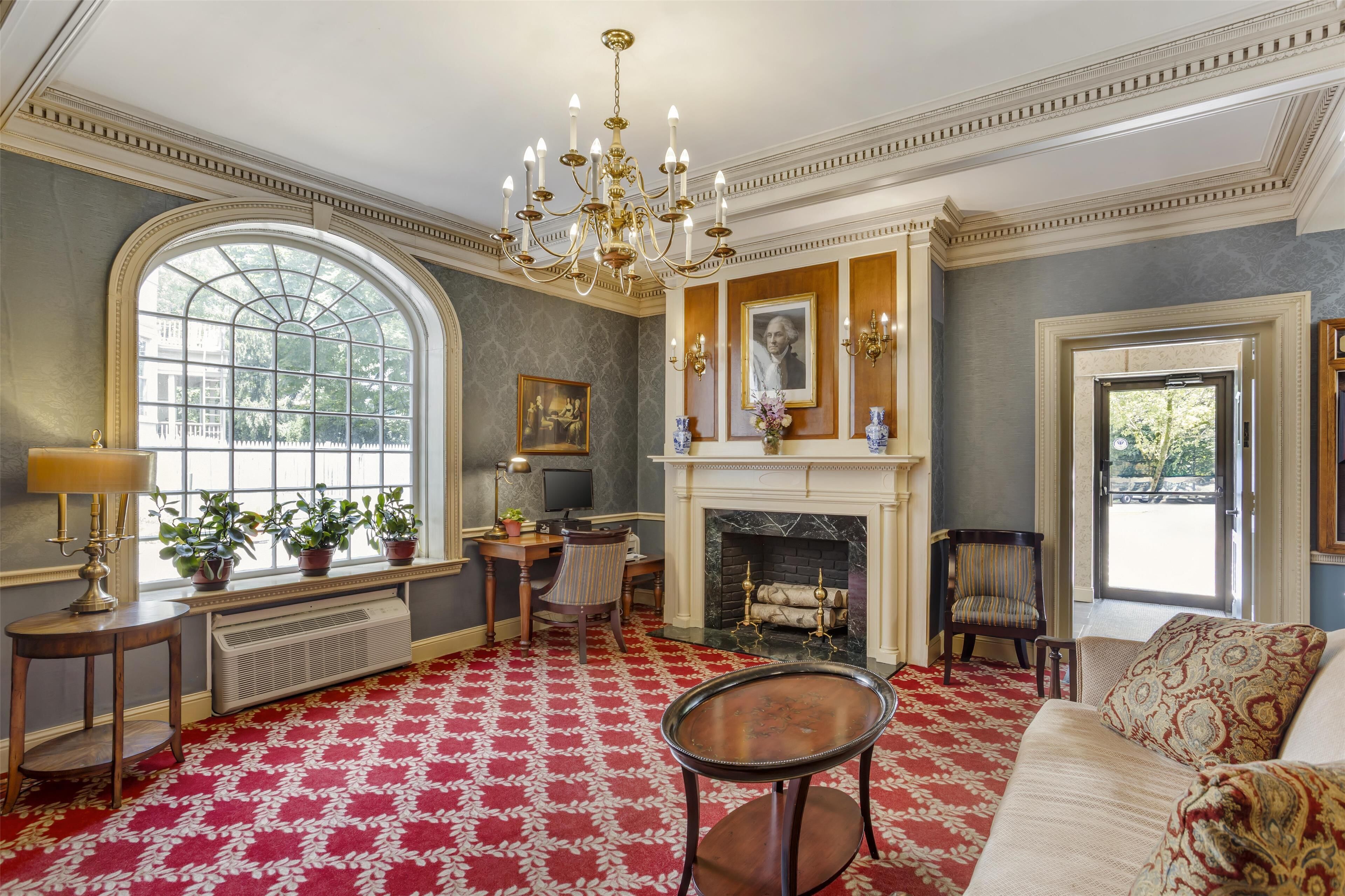 A room with a fireplace, a large window, a chandelier, and a red patterned carpet.