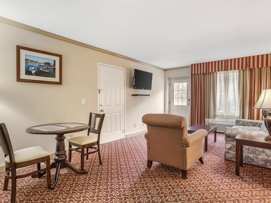 A hotel room with a dining table, two chairs, a sofa, a patterned carpet, a television, and a window with curtains.