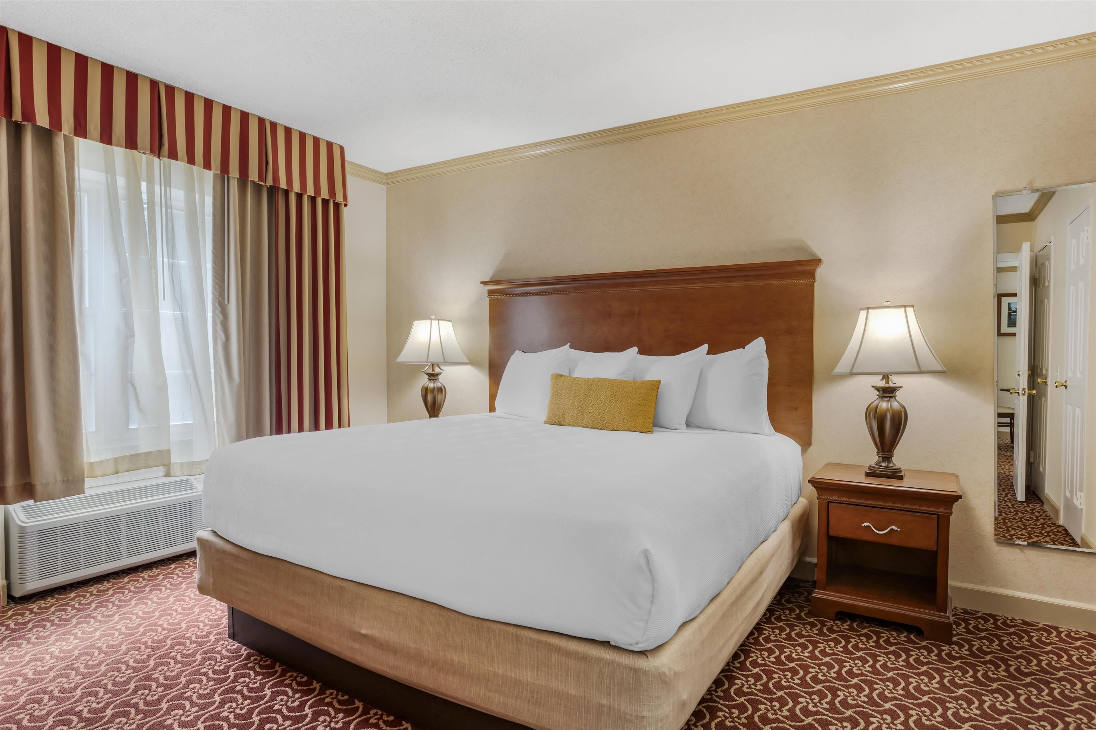 Habitación de hotel con una cama grande, dos mesitas de noche con lámparas, un espejo y una ventana con cortinas.