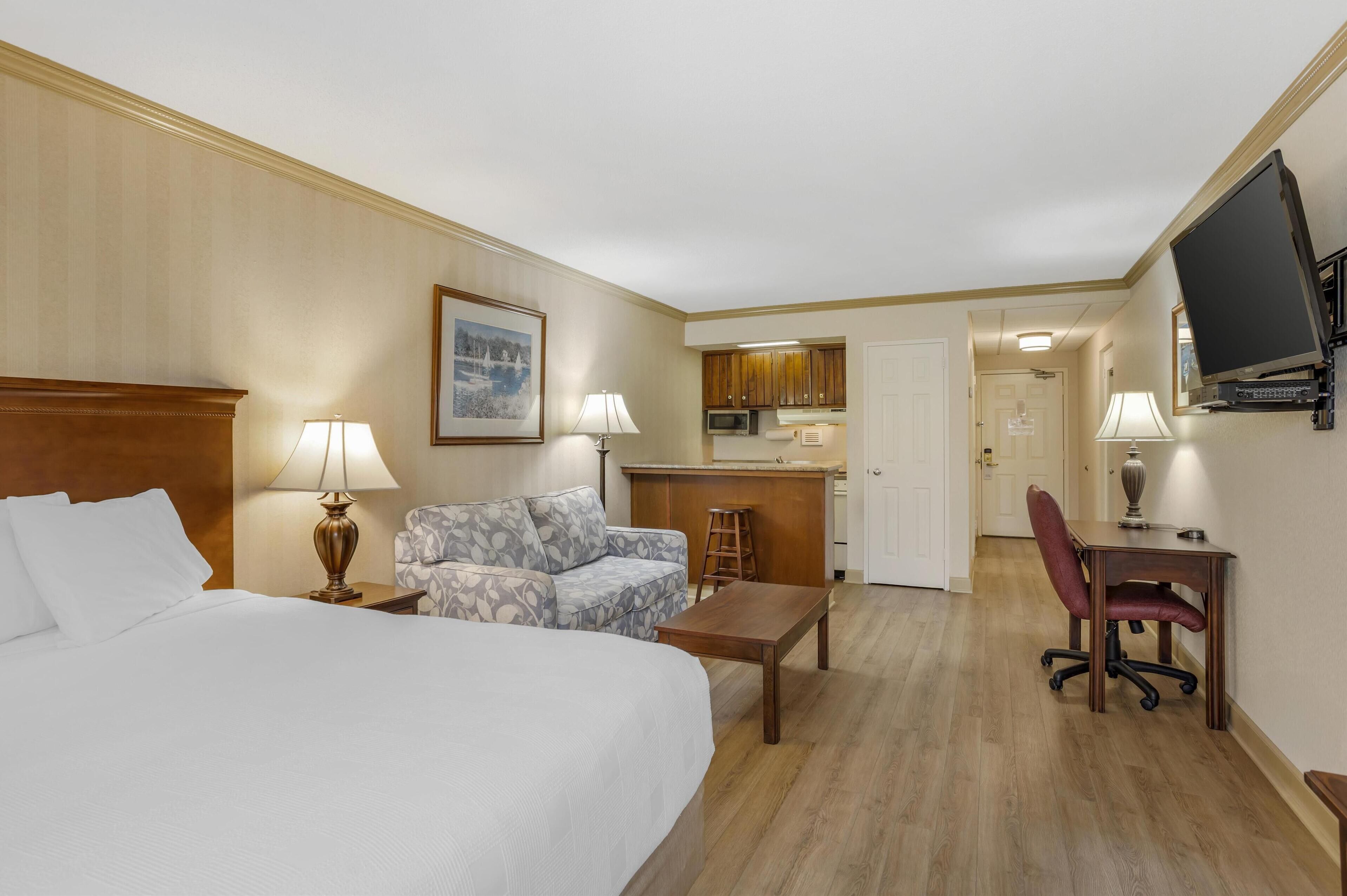 A hotel room with a large bed, a sofa, a desk with a chair, a television, a kitchen area, and a bathroom.