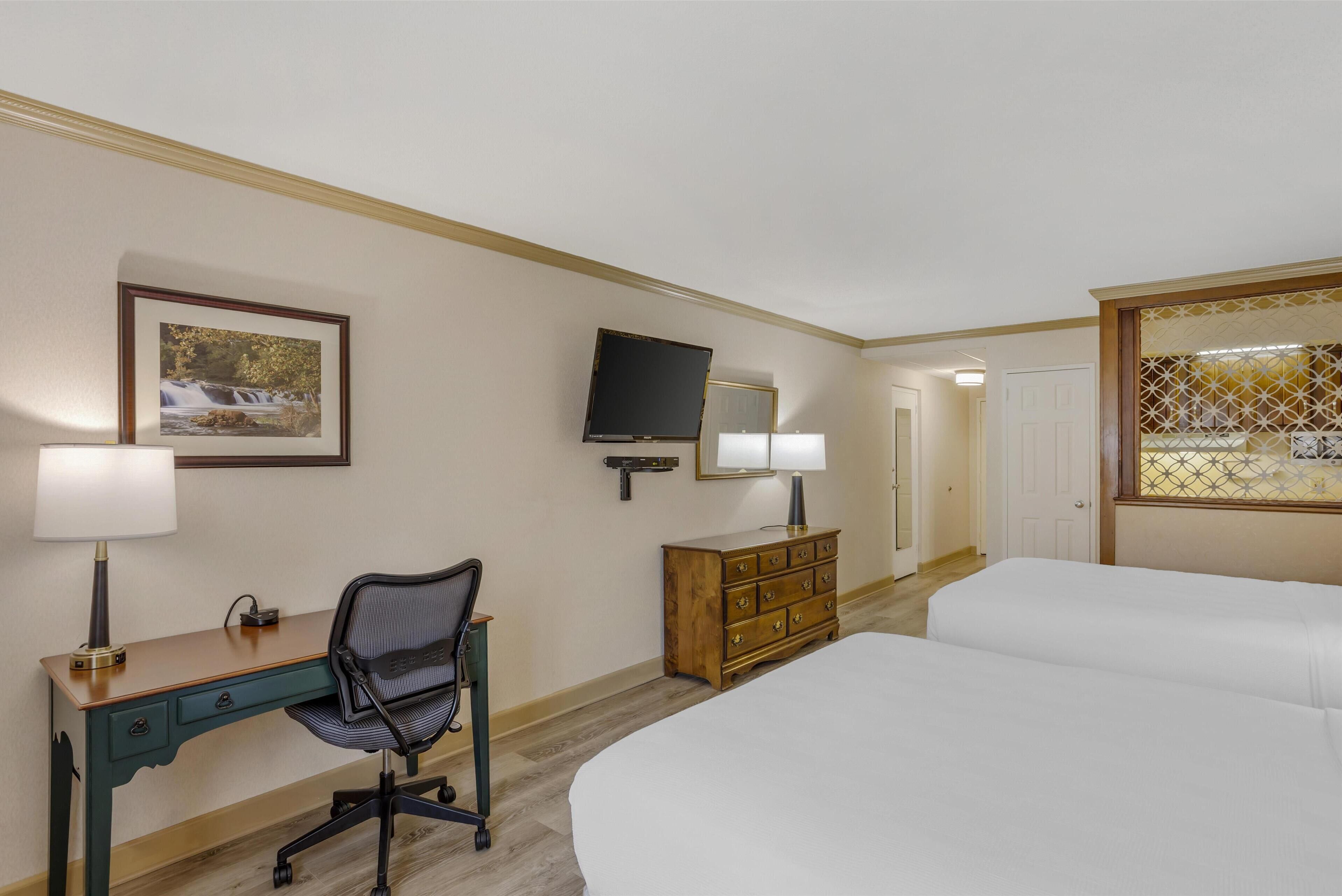A hotel room with a bed, a desk with a chair, a dresser, a television, and a framed picture on the wall.