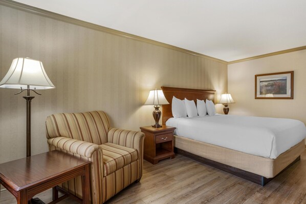 A hotel room with a bed, a striped armchair, a wooden nightstand with lamps, and a wooden table.