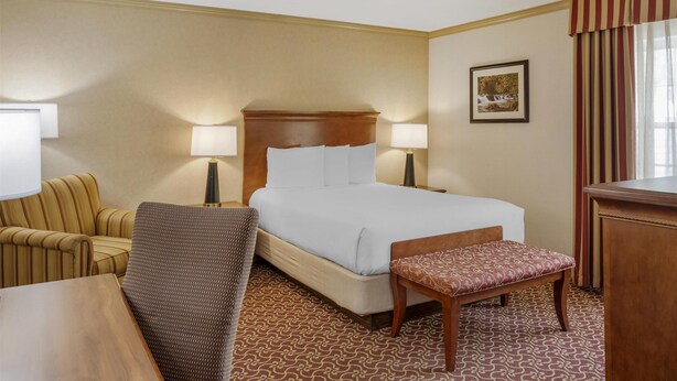 A hotel room with a bed, two bedside lamps, a chair, a bench, and a desk.