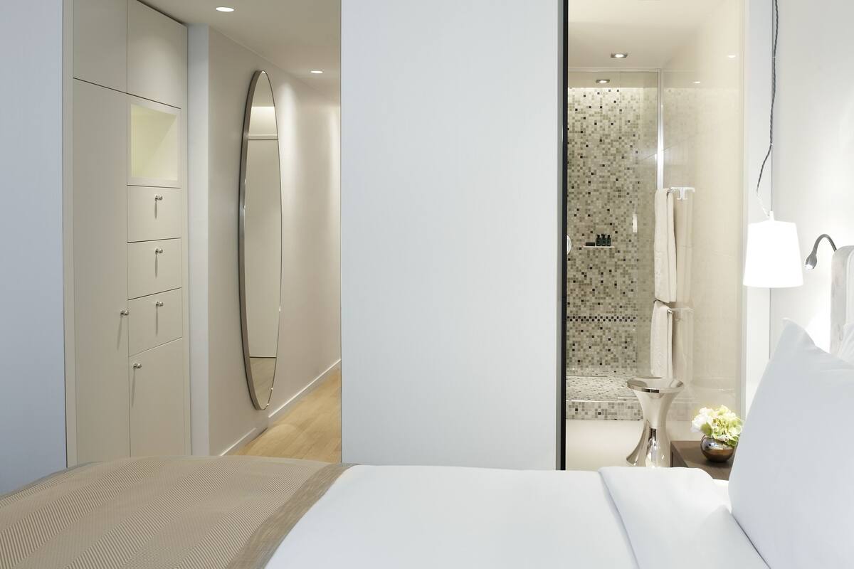 Una habitación de hotel moderna con una cama grande, un armario, un baño con ducha y un espejo.