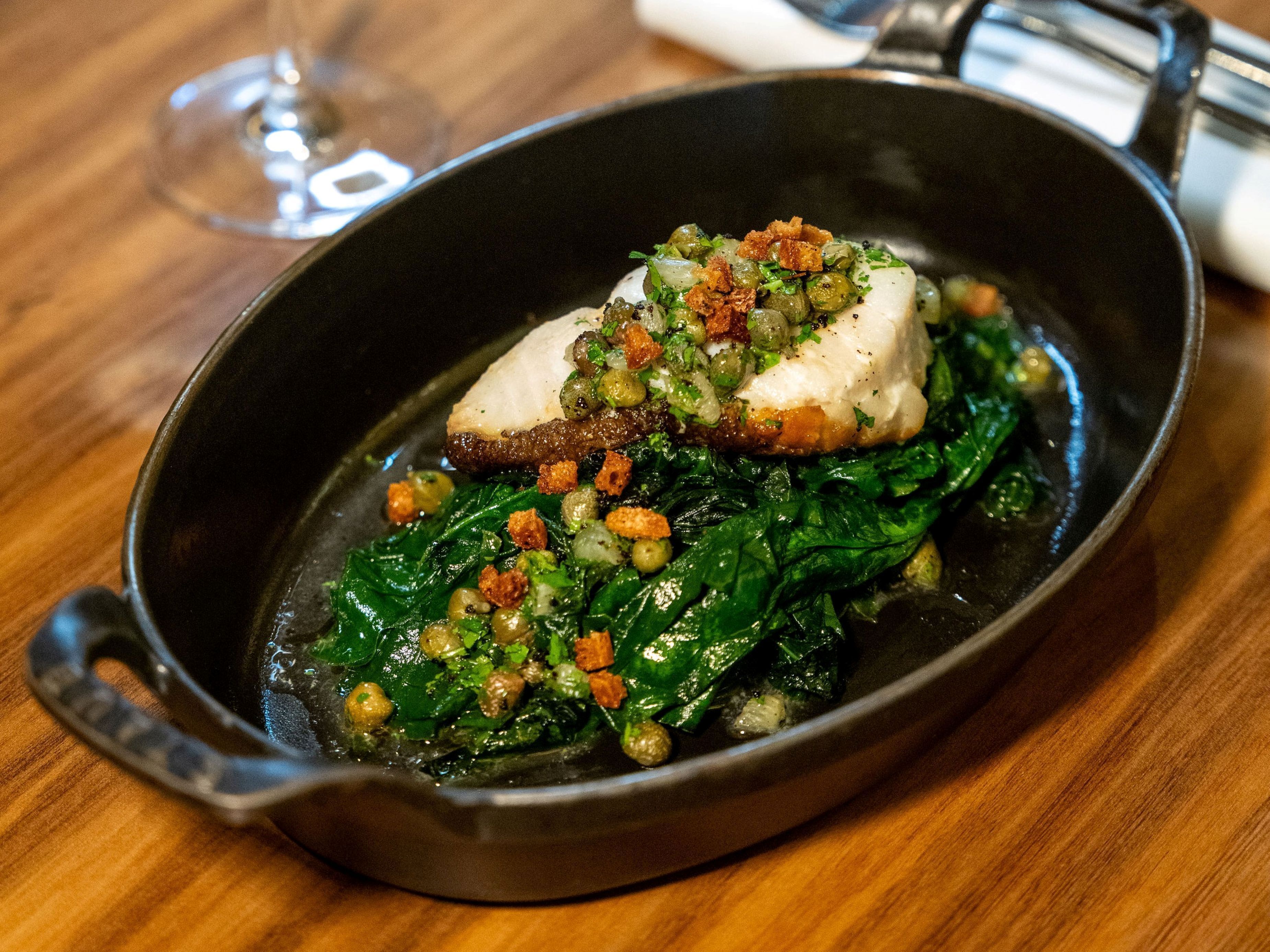 A skillet with a piece of fish and greens on a wooden table.
