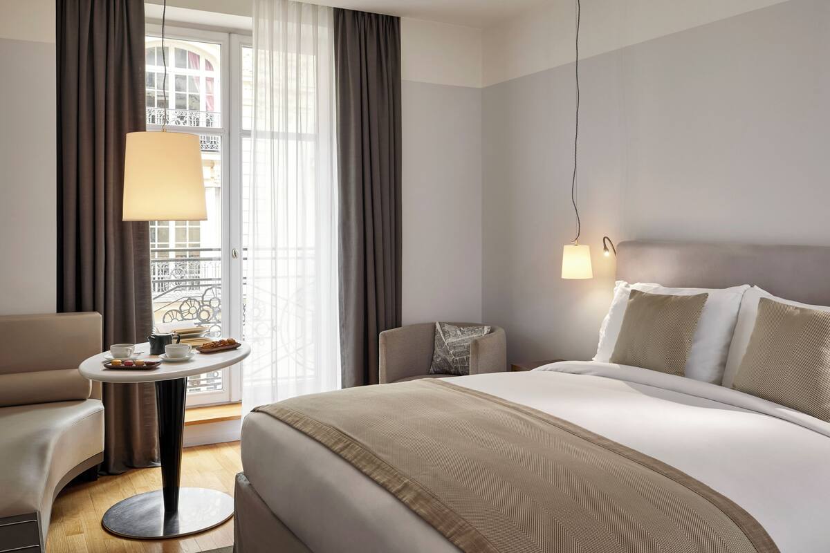 Una habitación de hotel con una cama, una mesa redonda con una lámpara, una silla y vistas a un balcón con cortinas.