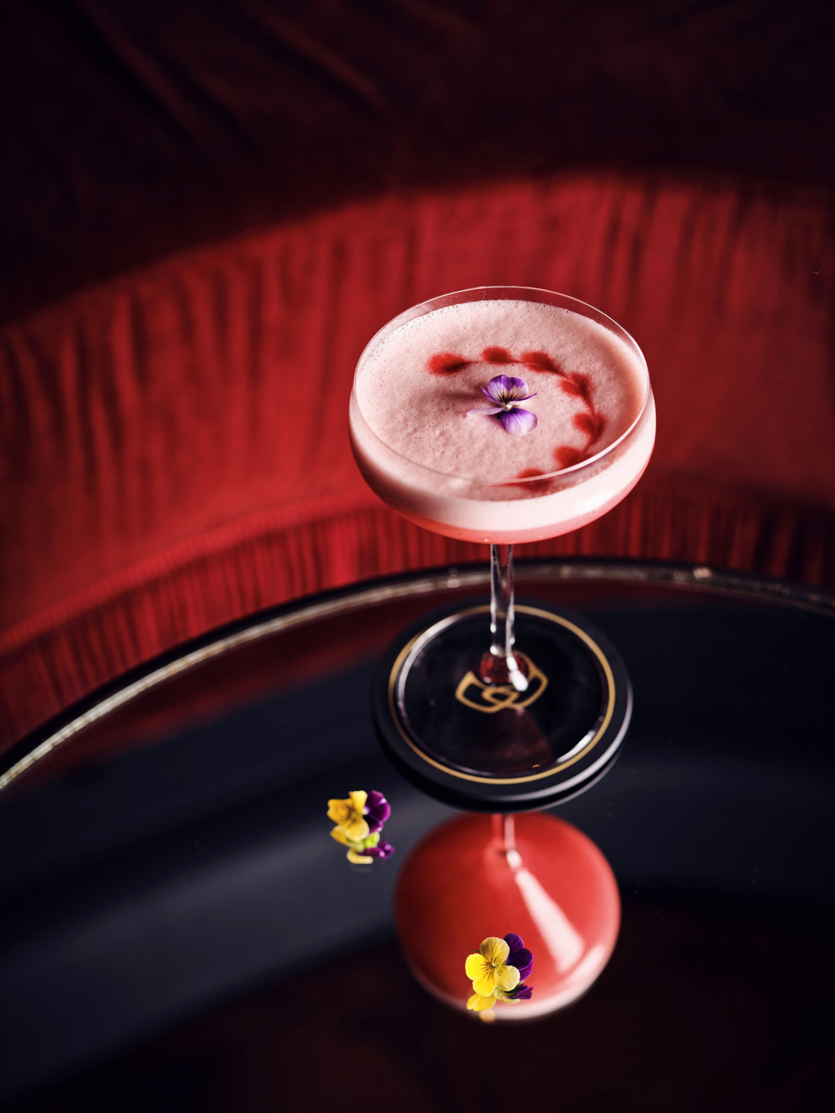 A cocktail in a glass with a flower garnish, placed on a reflective surface with a red background.