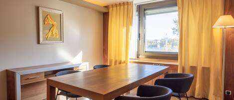 A modern room with a wooden dining table, two chairs, a desk with a chair, a lamp, and a window with curtains.