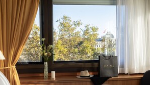 Una habitación de hotel con una cama, una ventana con cortinas, un florero con flores, una vela, un sombrero y un bolso en el alféizar.