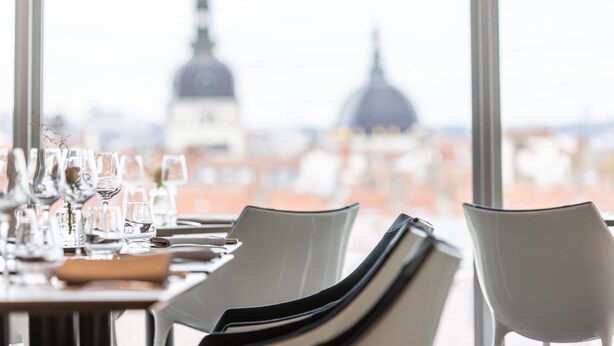 Mesa de comedor dispuesta con vasos y cubiertos, con vista a un paisaje urbano que incluye edificios con cúpulas.