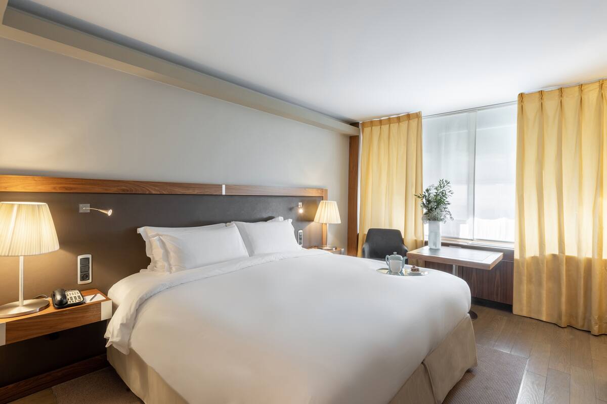 Hotelkamer met een groot bed, nachtlampjes, een telefoon op het nachtkastje, een klein tafeltje met een vaas en een raam met gordijnen.