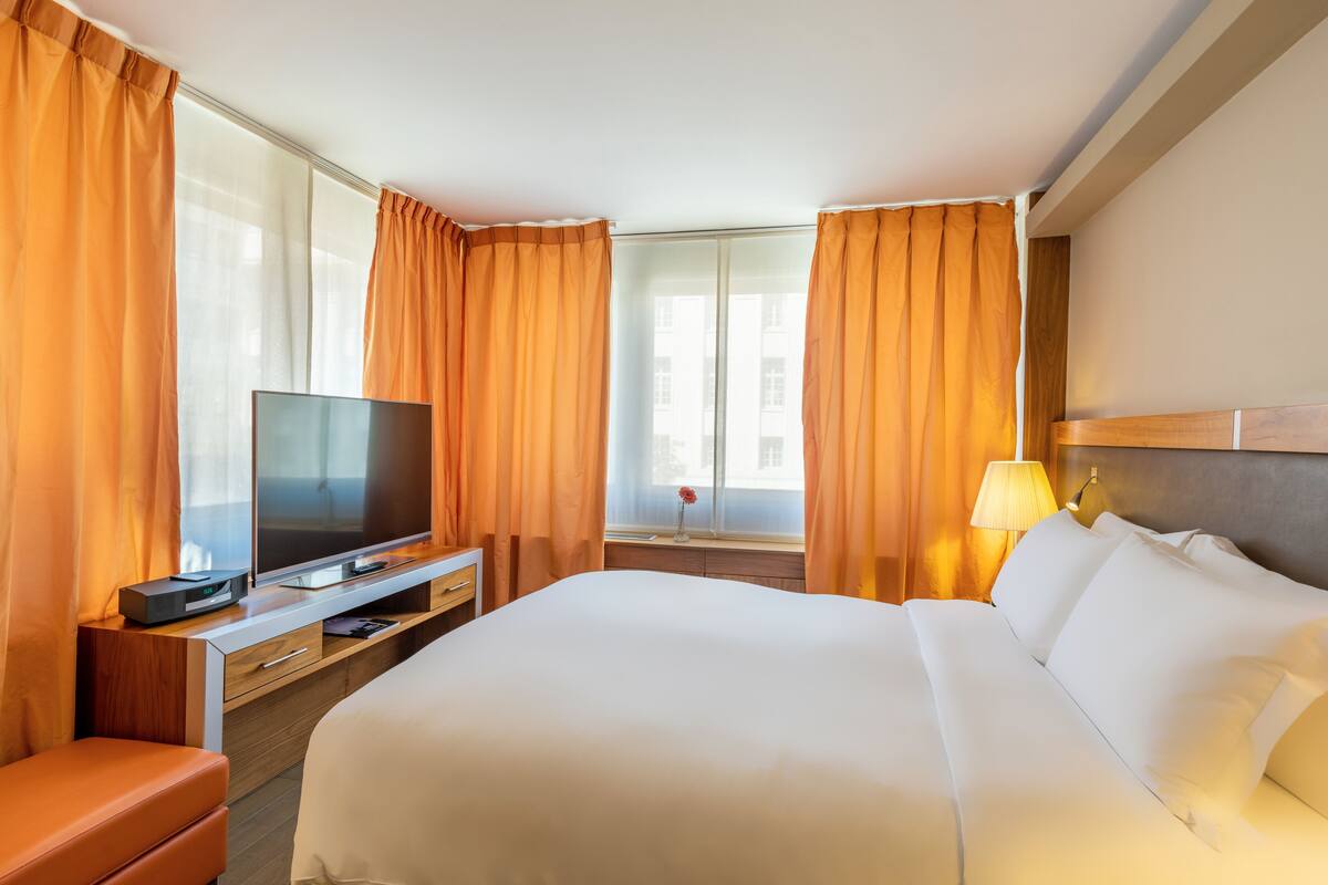 Een hotelkamer met een groot bed, een televisie op een houten dressoir, een lamp en oranje gordijnen.