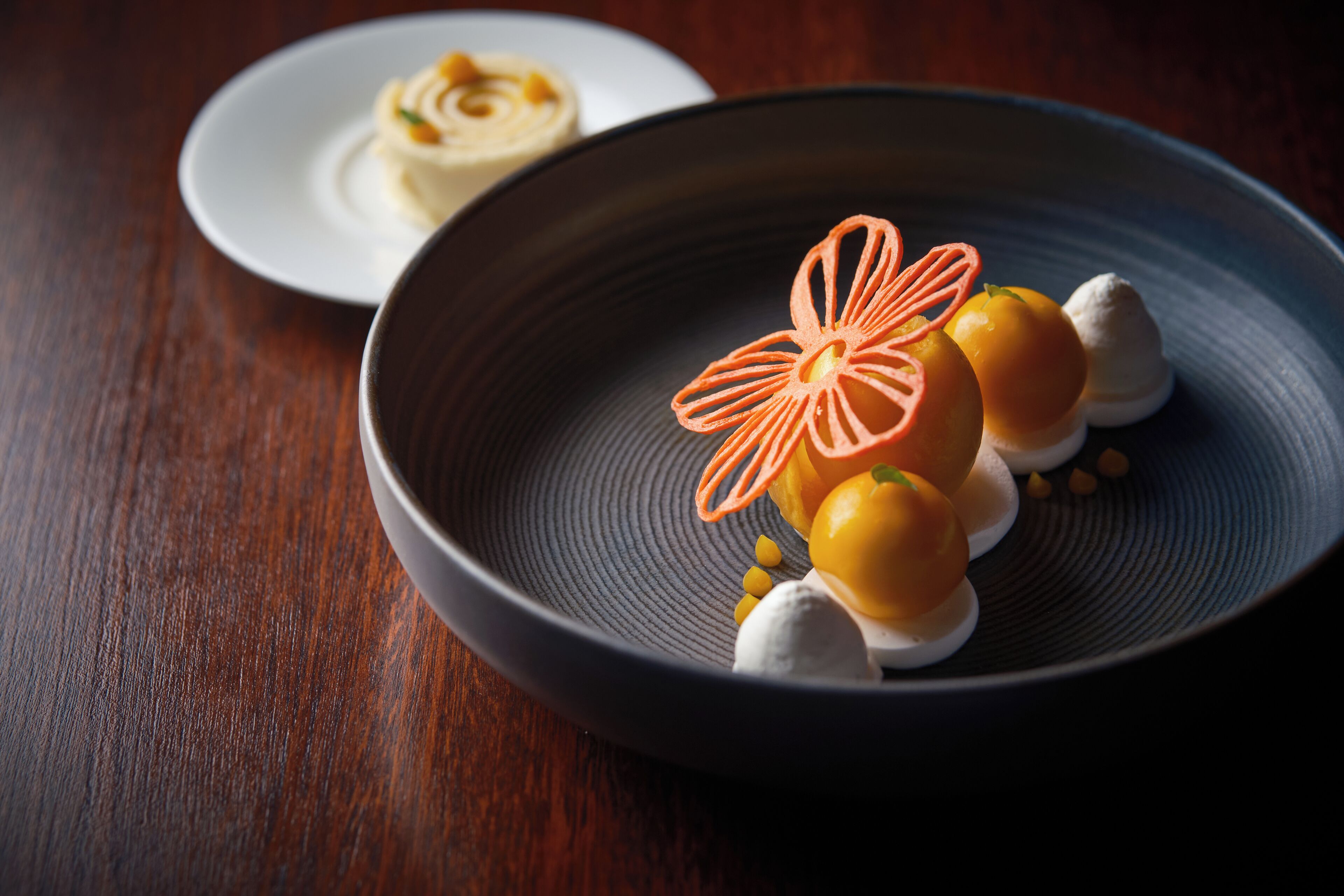 A dessert on a dark plate with a decorative orange flower, a yellow fruit, and small yellow seeds.