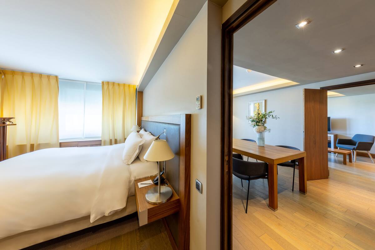 Een moderne hotelkamer met een groot bed, nachtkastje met lamp en uitzicht op de eetruimte.