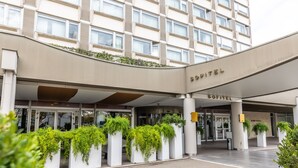 Entrada de un hotel con fachada curva, amplias ventanas y macetas alineando el camino.