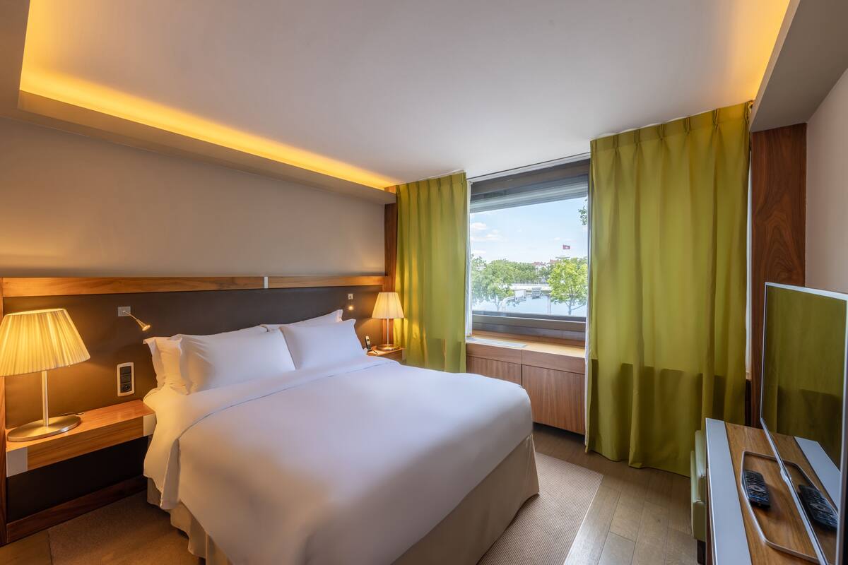 Een hotelkamer met een groot bed, nachtlampjes, een televisie en een raam met gordijnen.