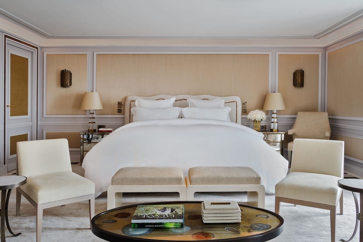 Une chambre d’hôtel avec un grand lit, deux fauteuils, une petite table avec un livre et une lampe, et une table de chevet avec une lampe et un vase de fleurs.