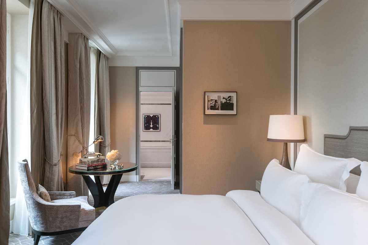 Une chambre d’hôtel avec un grand lit, une chaise, une petite table avec des livres et un vase, une lampe et un tableau encadré au mur.