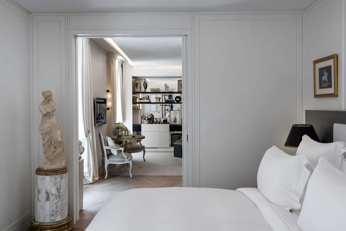 Une chambre d’hôtel avec une statue en marbre, un lit recouvert de draps blancs, une petite table avec des chaises et, au fond, un coin cuisine.