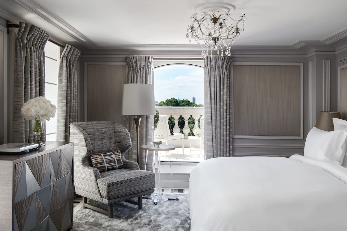 Une chambre d’hôtel dotée d’un grand lit, d’un coin salon avec un fauteuil et une table, d’un lustre et offrant une vue sur un balcon aménagé avec du mobilier d’extérieur.