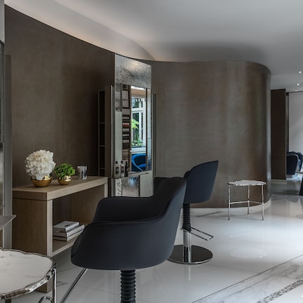 A modern salon with a curved wall, a marble countertop, and a large mirror. There are two black salon chairs, a small table, and a blue chair in the background.