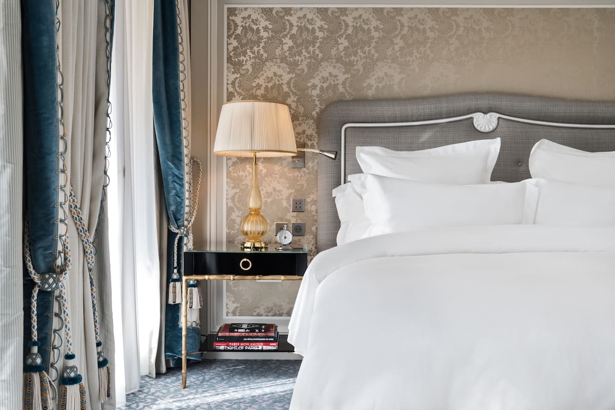 Un lit bien fait, avec du linge de lit blanc, une table de chevet avec une lampe et un mur recouvert d’un papier peint à motifs.