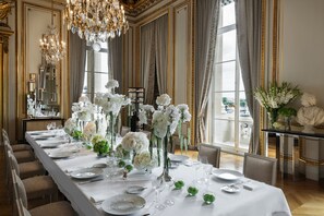 A formal dining room with a long table set for a meal, elegant chandeliers, and large windows with curtains.