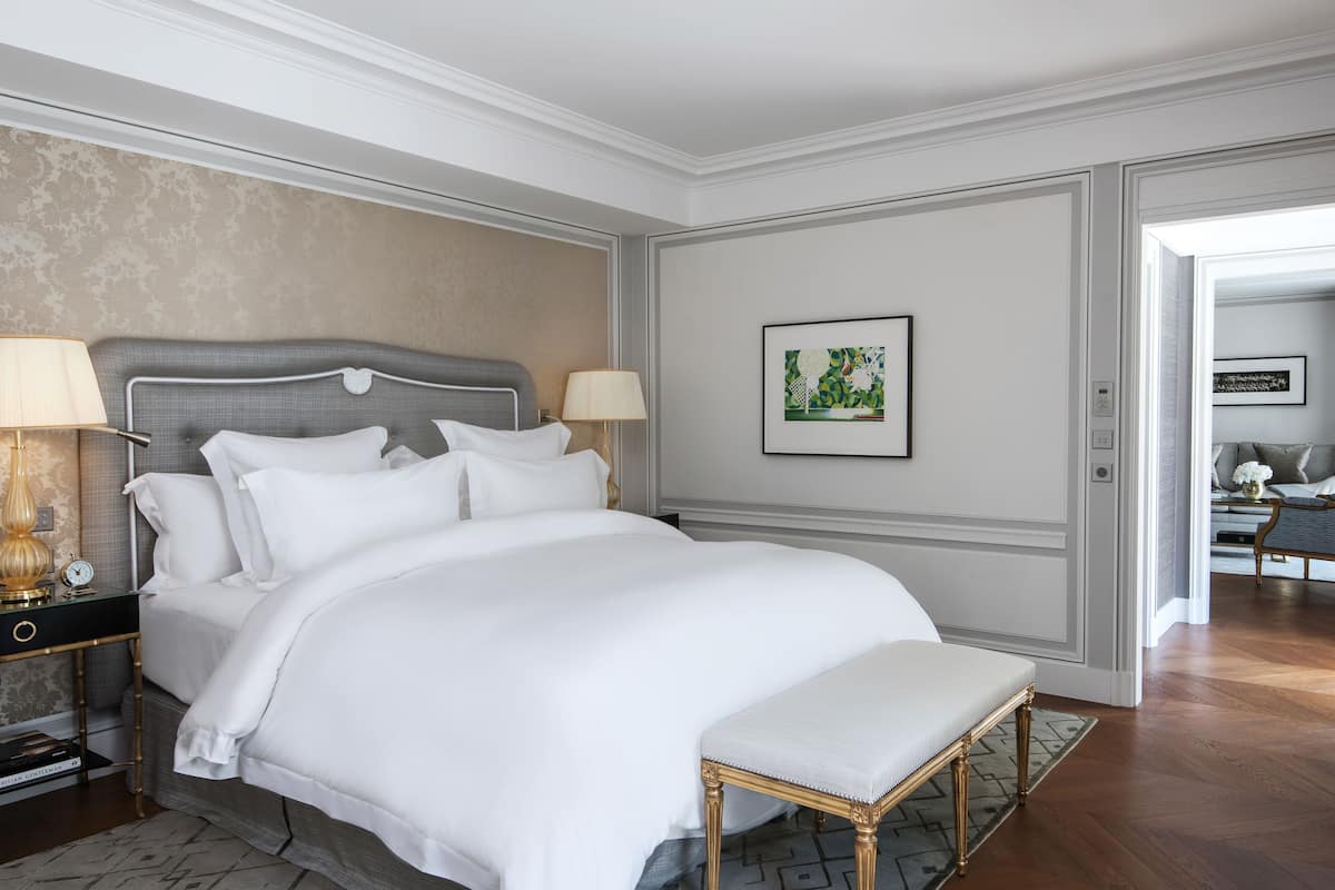Une chambre à coucher bien aménagée, avec un grand lit, des lampes de chevet, un banc et des œuvres d’art encadrées accrochées au mur.