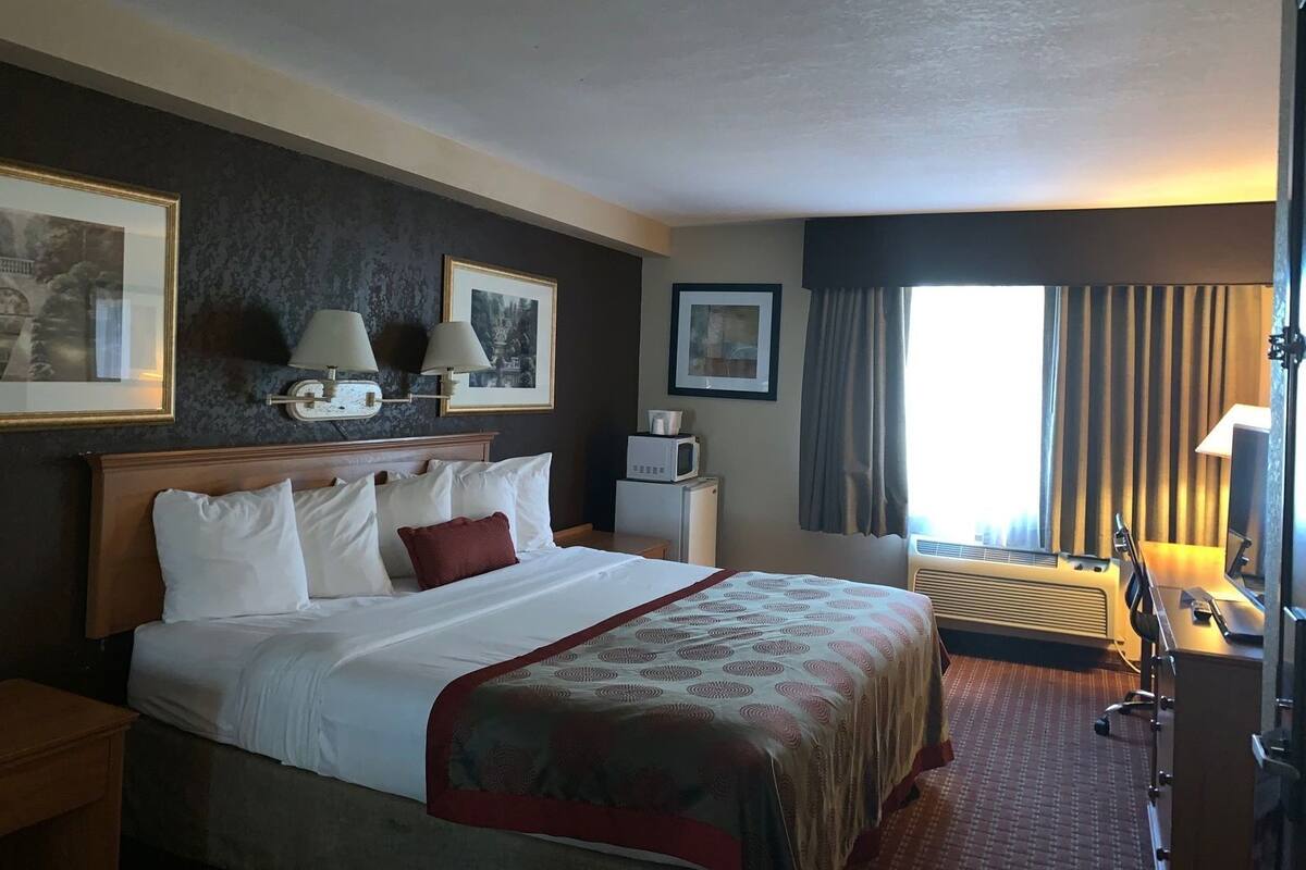 Ein Hotelzimmer mit Bett, Nachttischlampen, Schreibtisch, Stuhl, einem Fenster mit Vorhängen und Wandbildern.
