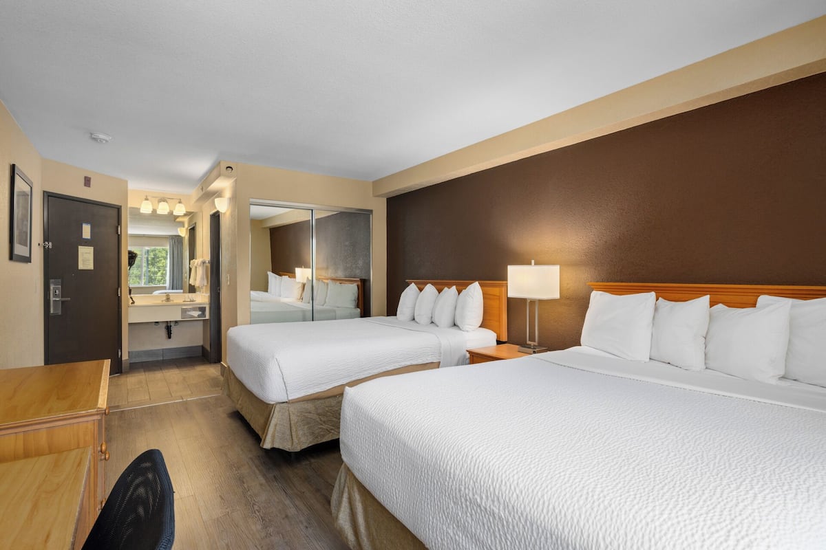 Ein Hotelzimmer mit zwei Betten, einem Schreibtisch, einem Stuhl, einem Spiegel, einem Badezimmer und einem Nachttisch mit Lampe.