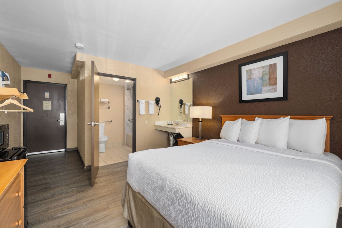 Ein Hotelzimmer mit einem großen Bett, einem Schreibtisch mit Computer, einem Badezimmer mit Dusche und einer Tür zu einem weiteren Raum.