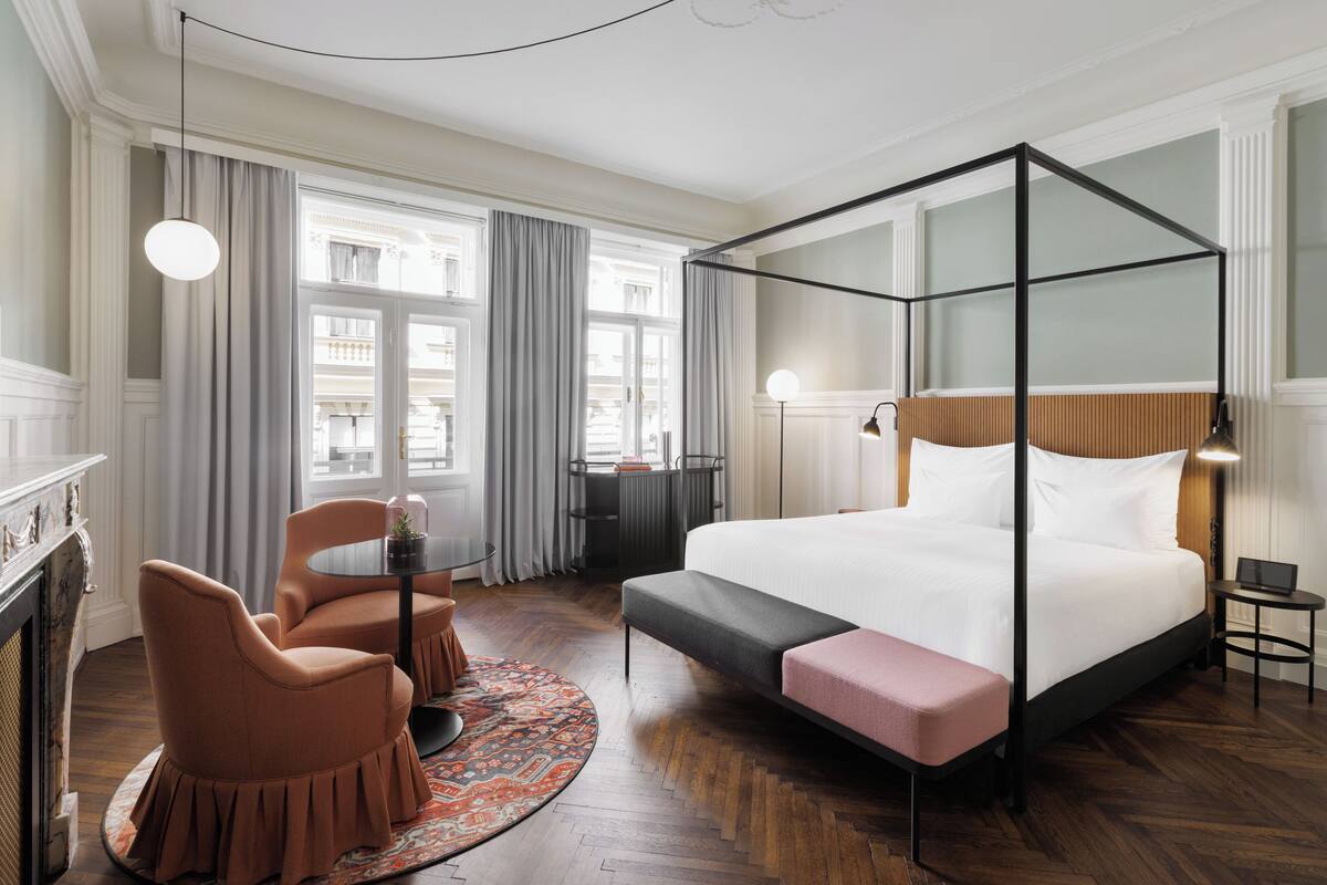 A modern hotel room with a four-poster bed, a round table with chairs, a fireplace, and a view of the city.