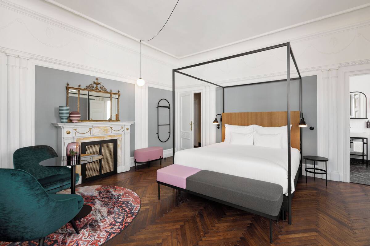 A modern hotel room with a four-poster bed, a fireplace, a sitting area with a chair and a small table, and a bathroom visible through an open door.