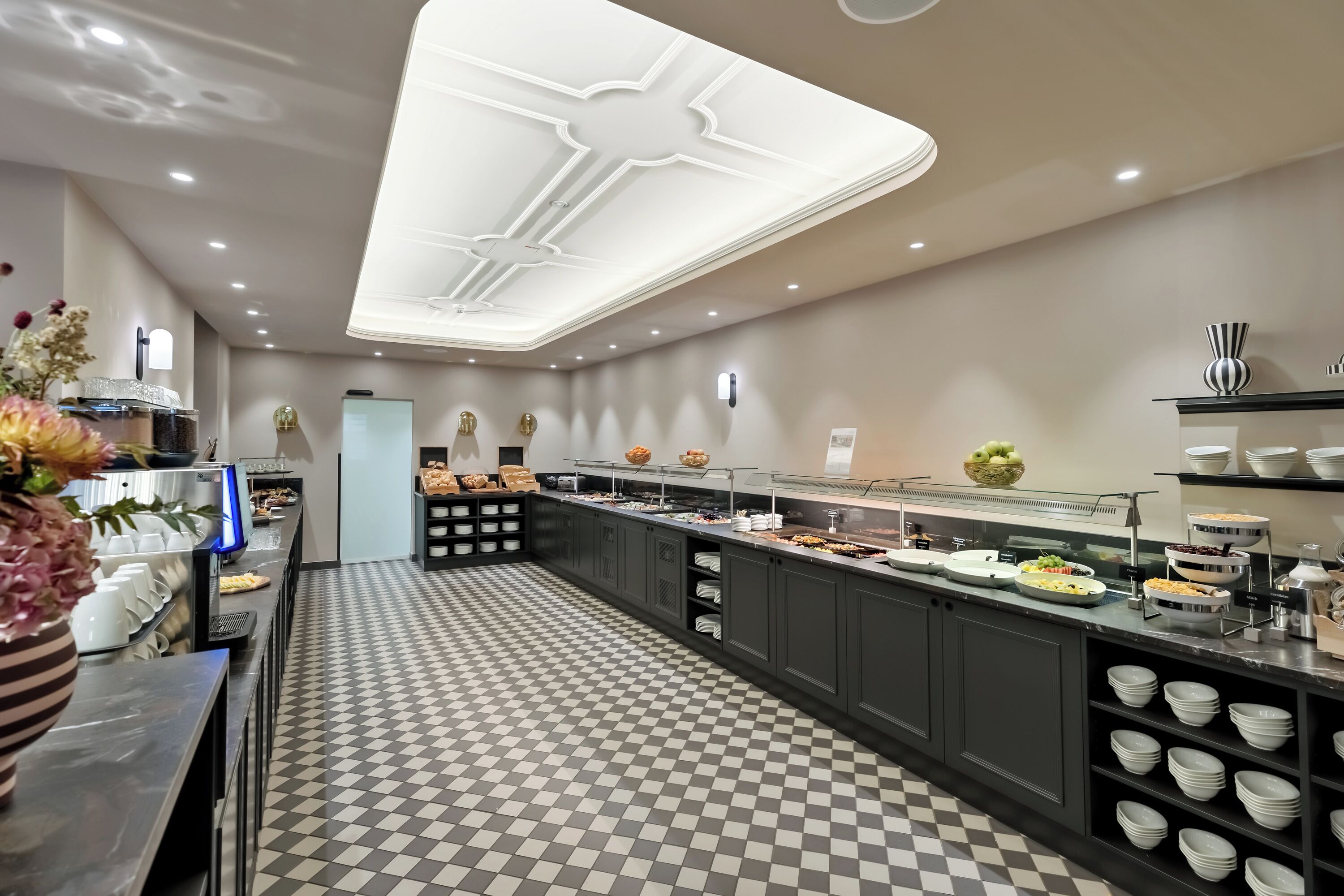 A modern hotel lobby with a checkered floor, a central reception desk, and a buffet area with various food options.