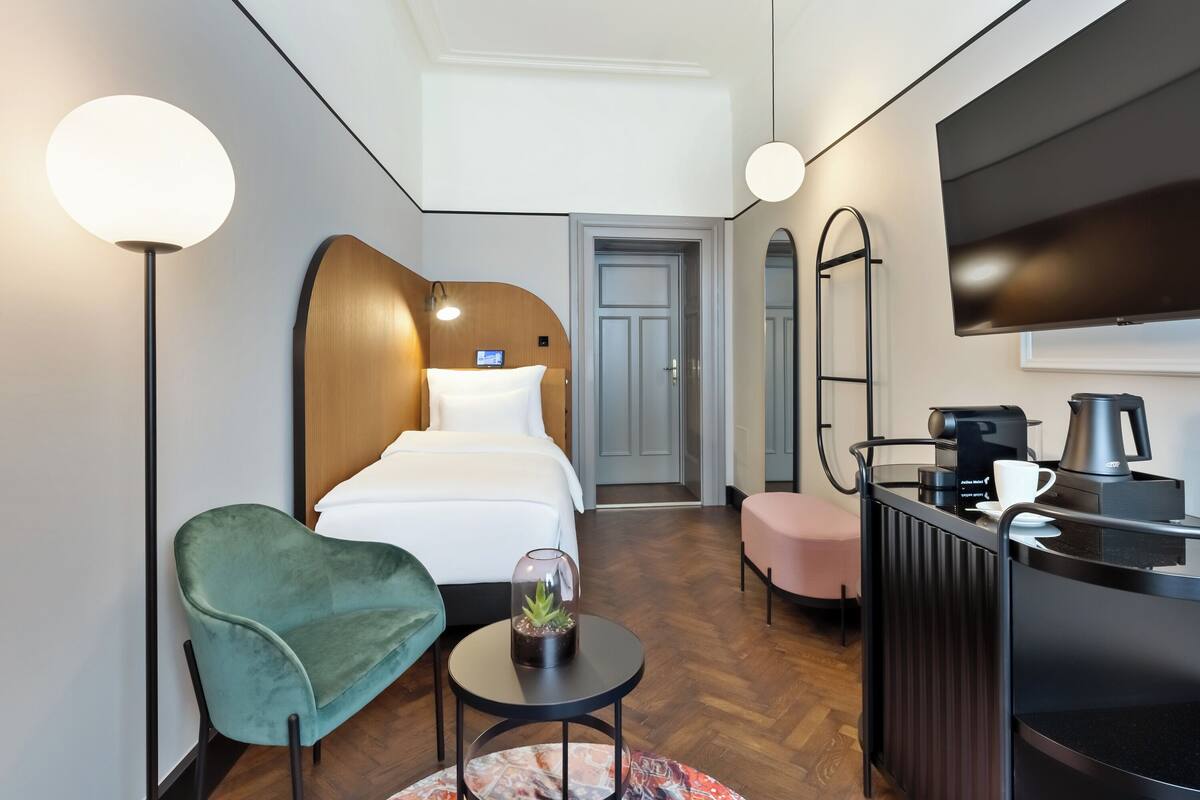 A modern hotel room with a bed, a green armchair, a small table with a plant, a coffee machine, and a TV mounted on the wall.
