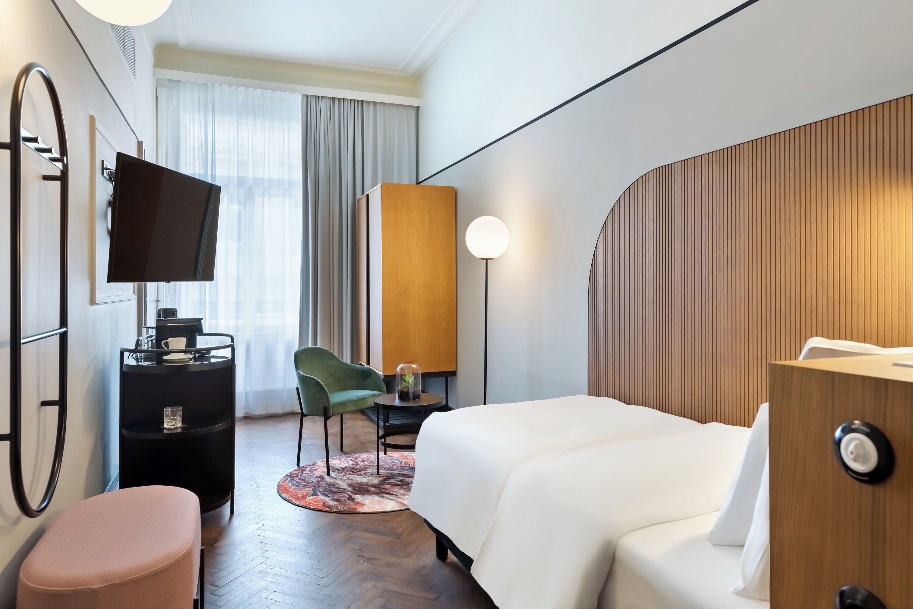 A hotel room with a bed, a desk with a computer, a chair, a TV, and a window with curtains.