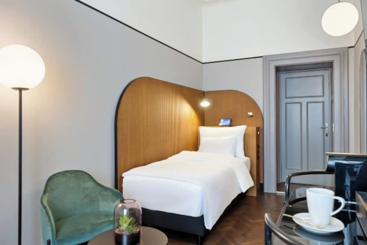 A hotel room with a bed, a desk with a chair, a lamp, and a door.