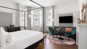 Una habitación de hotel moderna con una cama grande, un sofá, un televisor y vistas a la ciudad a través de amplios ventanales.