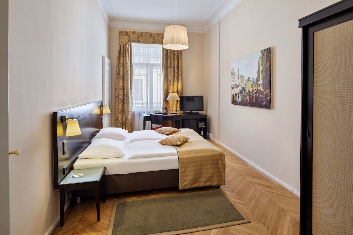 Quarto de hotel com cama, abajures, televisão, pintura e janela com cortinas.