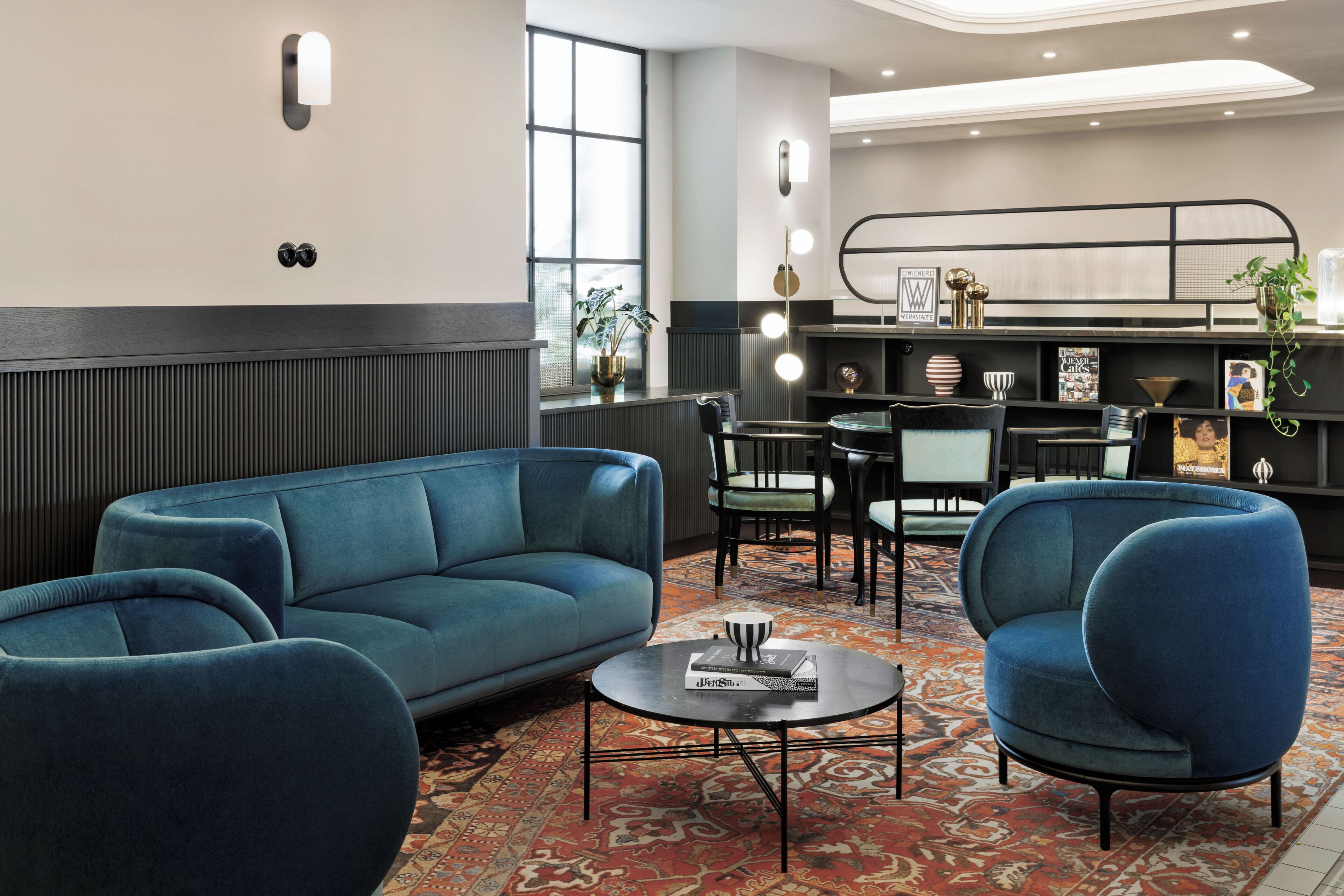 Un área de estar moderna con un sofá azul, sillones, una mesa redonda de centro y una alfombra estampada.