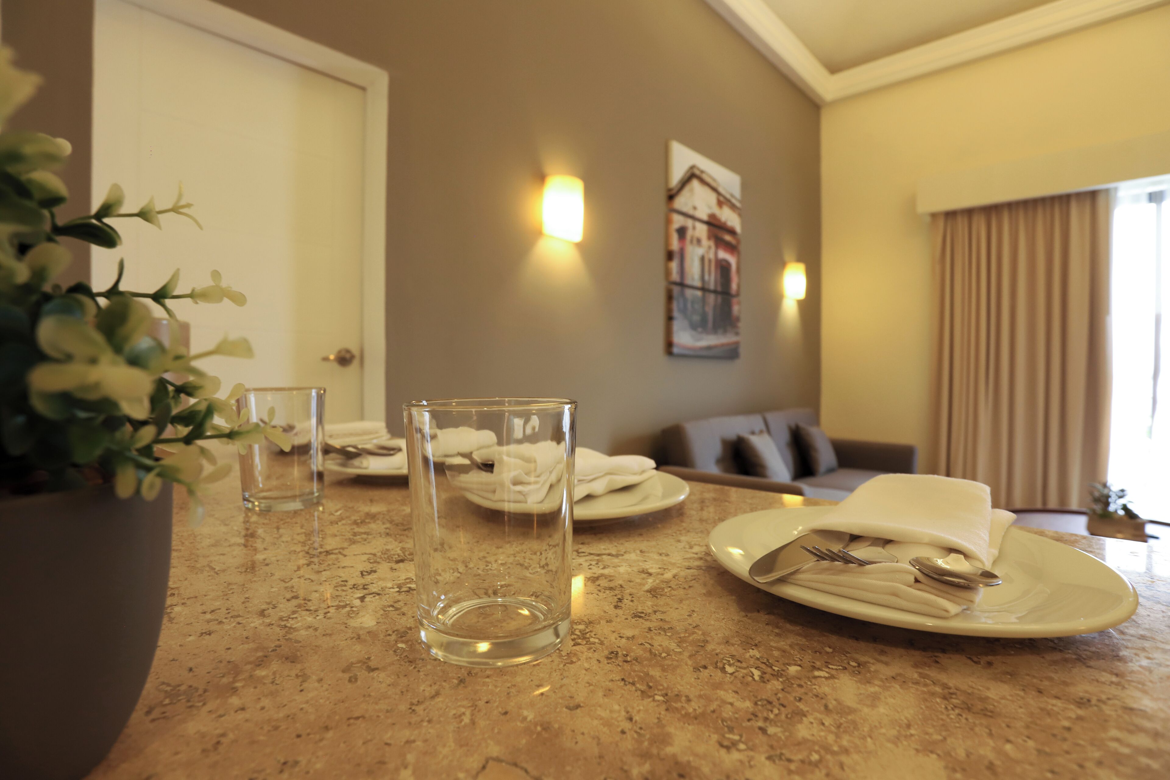 A hotel room with a dining table set for two, a potted plant, and a painting on the wall.