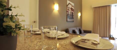 A hotel room with a dining table set for two, a potted plant, and a painting on the wall.
