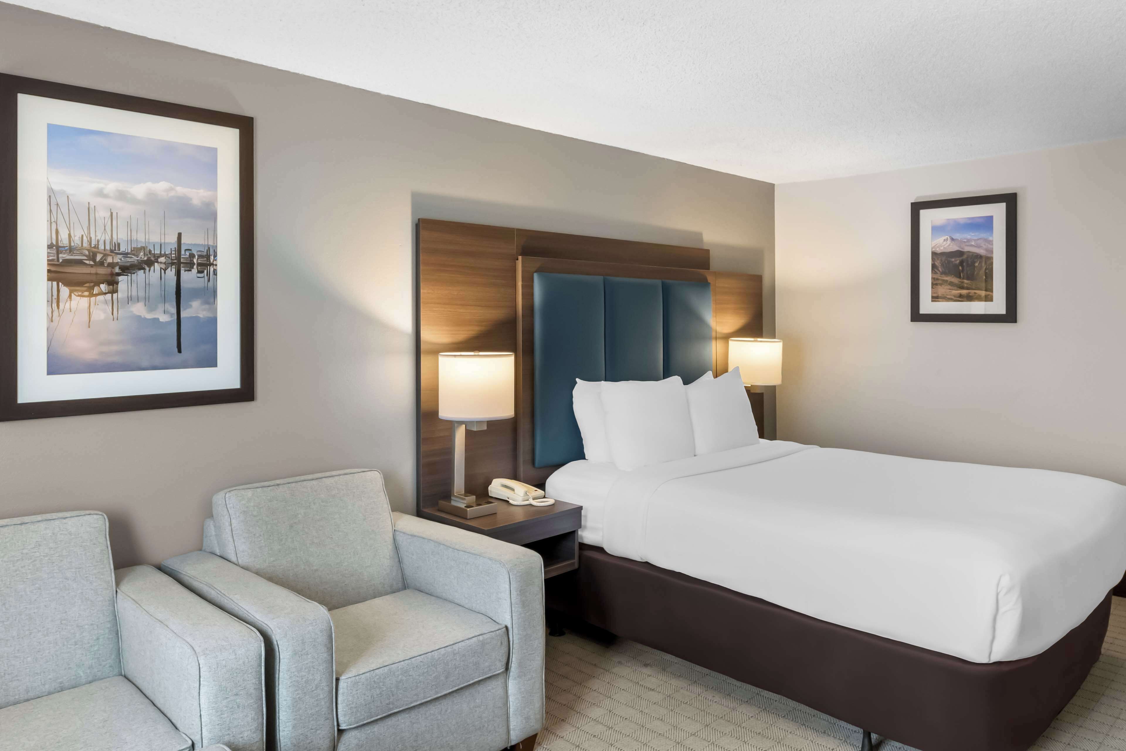 A hotel room with a large bed, a desk with a lamp, a chair, and two framed pictures on the walls.