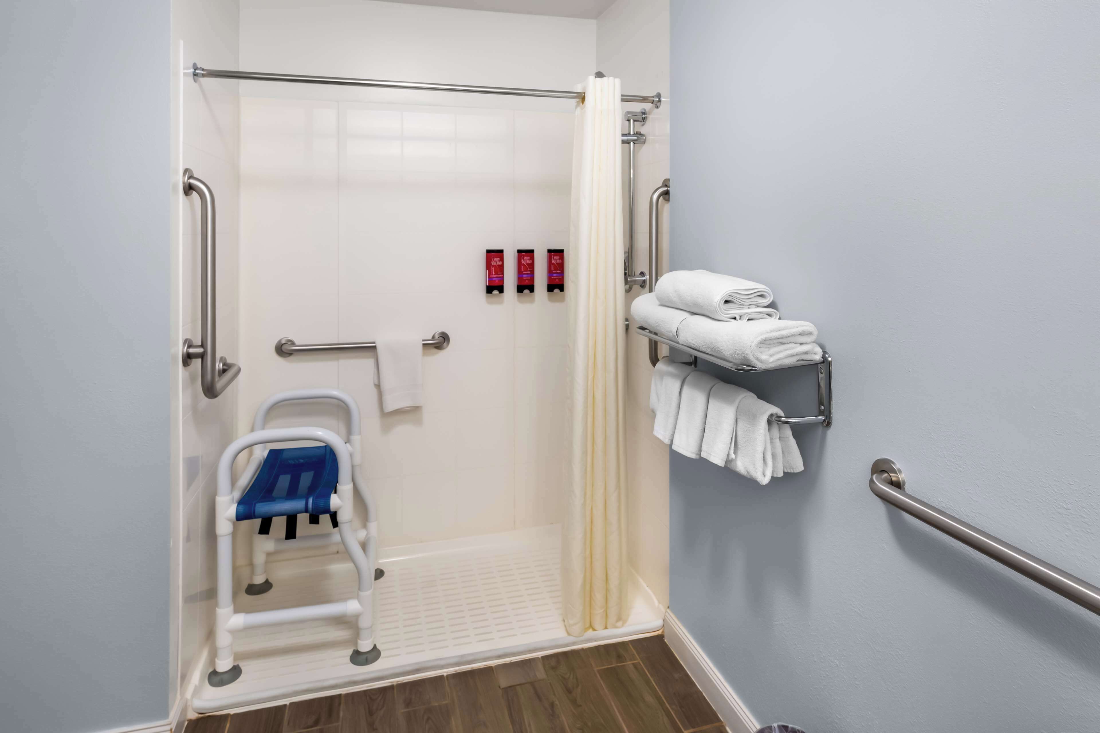 A bathroom with a shower, grab bars, a wheelchair-accessible bench, and a towel rack with folded towels.