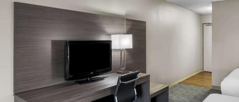 A hotel room with a flat-screen TV, a desk with a lamp, a black office chair, and a bed.