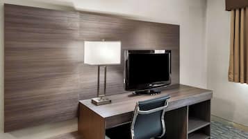 A modern hotel room with a wooden desk, a flat-screen TV, a lamp, and a chair.
