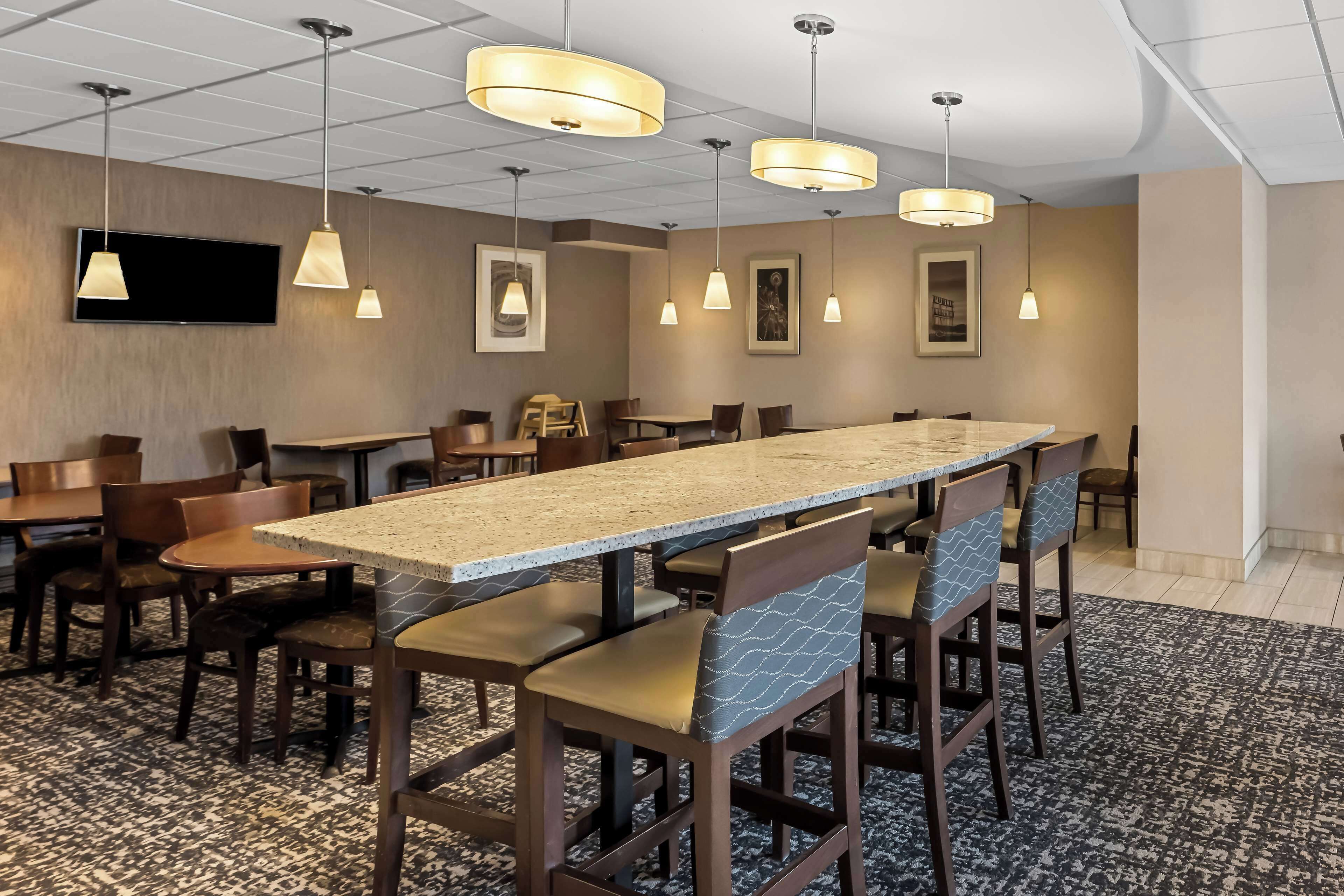 A hotel dining area with a long communal table, multiple chairs, hanging pendant lights, and framed artwork on the walls.