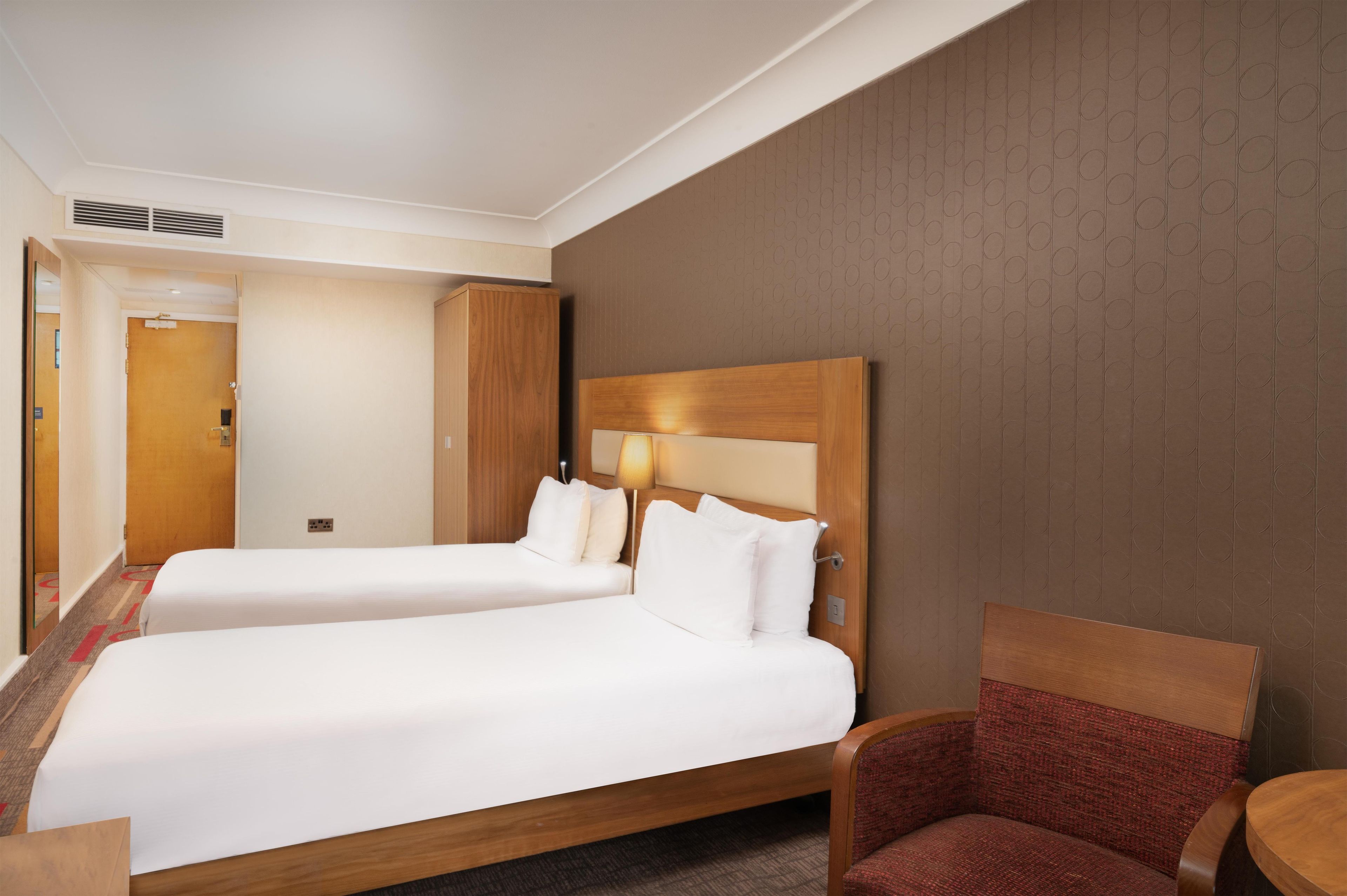 Habitación de hotel con dos camas, un cabecero de madera, un sillón tapizado en rojo y suelo alfombrado.