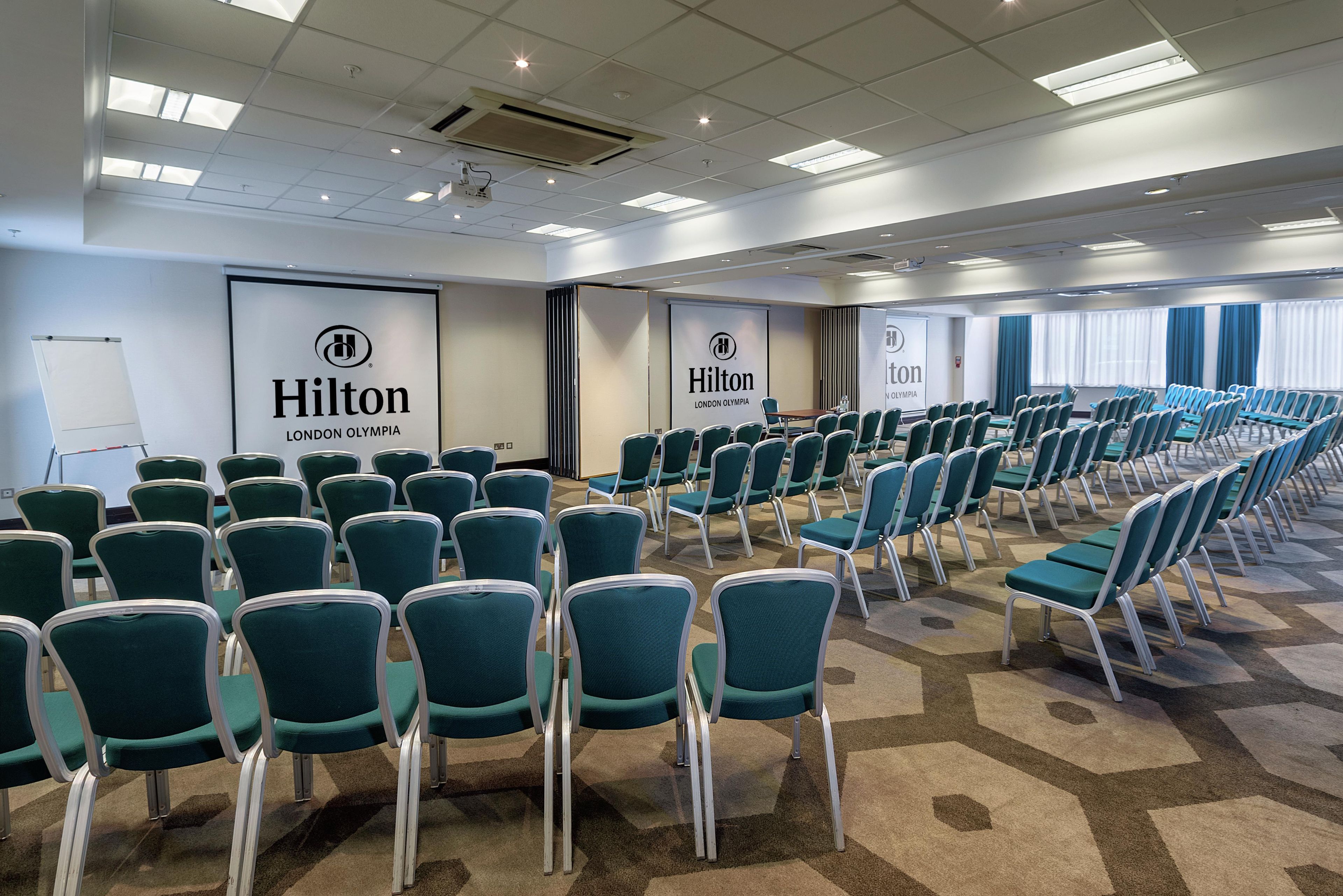 A conference room with rows of teal chairs, a whiteboard, and Hilton branding on the walls.