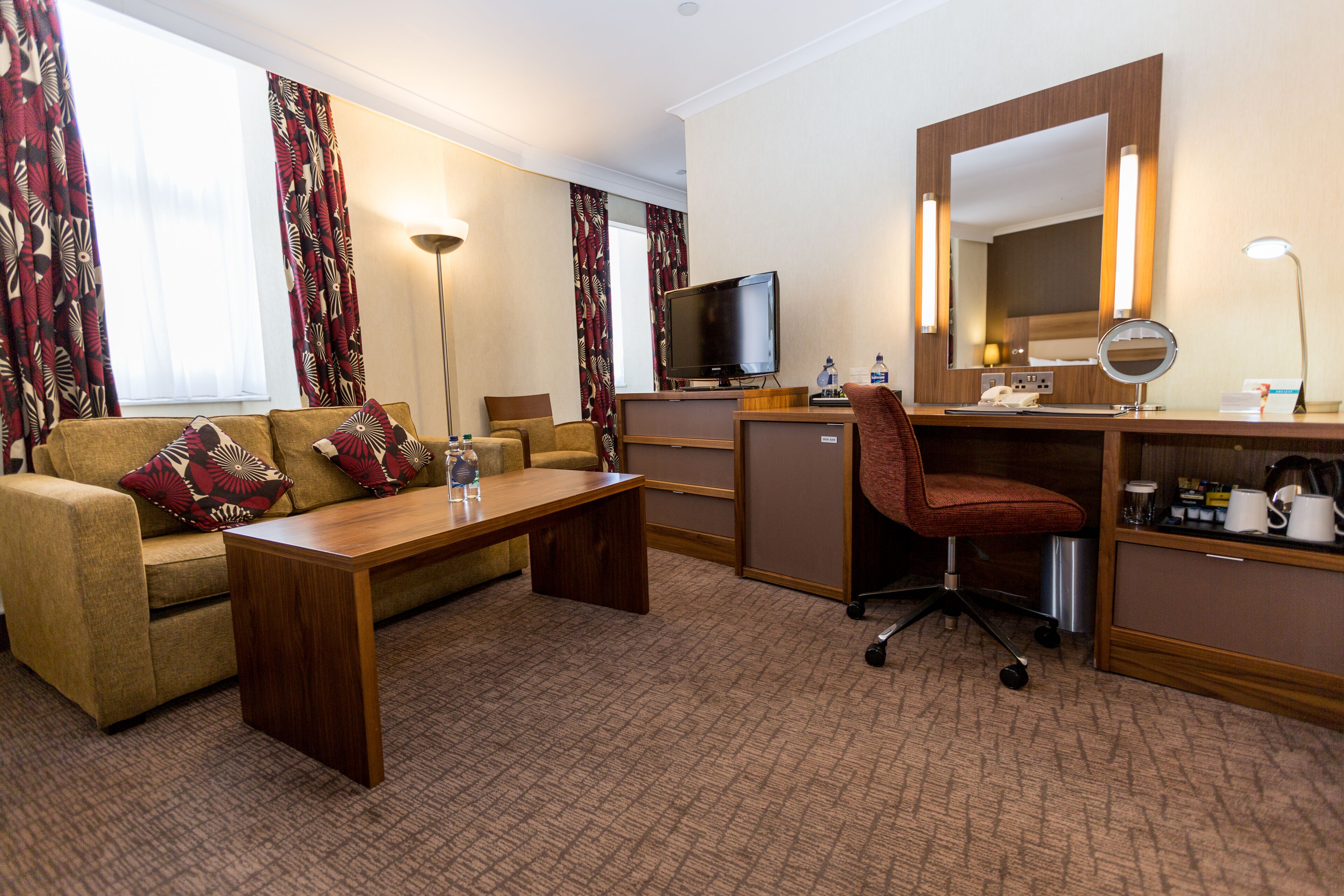A hotel room with a sofa, a wooden coffee table, a desk with a computer and a mirror, a chair, and a television.